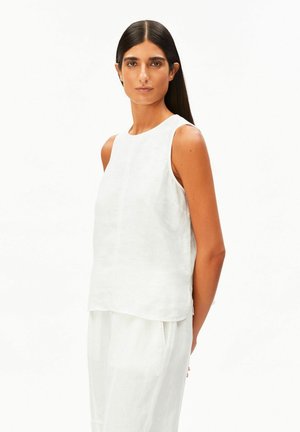 Frau mit langen dunklen Haaren, die ein ärmelloses, lockeres weißes Oberteil und passende weiße Hose trägt, posiert vor einem schlichten weißen Hintergrund.