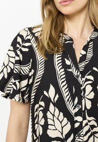 Camicia nera e crema con motivo di foglie botaniche, maniche corte a volant, chiusura con bottoni e tessuto testurizzato.
