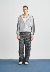 Γκρι φούτερ με φερμουάρ και μαύρες ρίγες, σε συνδυασμό με άνετο παντελόνι σε μαύρο και γκρι με κυματιστό σχέδιο. Ολοκληρώνεται με λευκά αθλητικά παπούτσια.