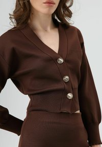 Bruin, geribd gebreid cropped topje met V-hals, voorzien van drie grote zilveren knopen. Lange mouwen met aansluitende manchetten. Gladde textuur.