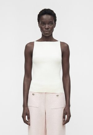 Femme aux cheveux courts et bouclés portant un haut blanc sans manches et un pantalon taille haute rose clair avec poches devant, sur un fond uni.