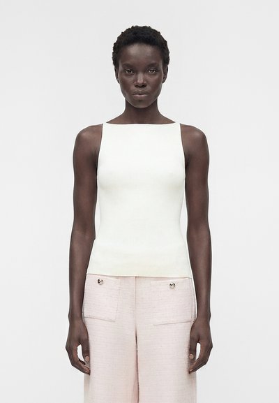 Femme aux cheveux courts et bouclés portant un haut blanc sans manches et un pantalon taille haute rose clair avec poches devant, sur un fond uni.