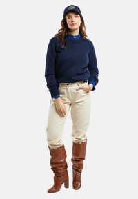 Pull marin, chemise bleue à col, pantalon crème et bottes en daim marron hautes à la cheville. Comprend une casquette avec logo et une coupe décontractée.