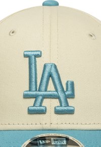 Gorra de béisbol beige con el logo "LA" bordado en azul claro en la parte frontal. La tela tiene una textura con un acabado liso y estructurado.