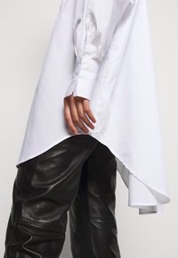 Persona que lleva una camiseta larga y asimétrica blanca con puños abotonados y pantalones de cuero negros, con la mano relajada a su lado.