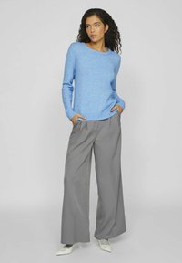 Mėlynas megztinis su ilgomis rankovėmis ir banguotais rankogaliais, derinamas su šviesiai pilkomis klostuotomis plačiomis kelnių, turinčiomis vertikalias juostas.