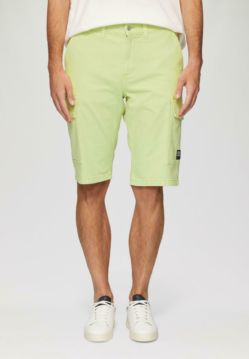 Jasnozielone szorty cargo z kieszeniami bocznymi, zapięciem na guziki i podwiniętymi nogawkami. W komplecie z białymi trampkami i beżową koszulą.