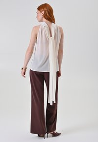 Blusa bianca senza maniche con un legame in tessuto drappeggiato sul retro, abbinata a pantaloni larghi marroni. La blusa ha una texture trasparente.