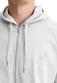 Grå zip-up hoodie med huva, som har dragsnöre, en diskret logo på bröstet och ribbad textur vid halsringningen och ärmsluten.