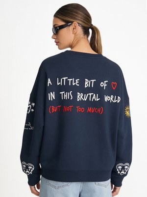 Femme avec une queue de cheval brune et des lunettes de soleil porte un sweat-shirt bleu marine avec le texte : "A little bit of love in this brutal world (but not too much)."