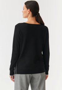 Schwarzer Pullover mit weitem Ausschnitt, gerippten Bündchen und seitlichen Schlitzen. Über grauen Hosen getragen, die eine glatte Textur und ein lässiges Design präsentieren.