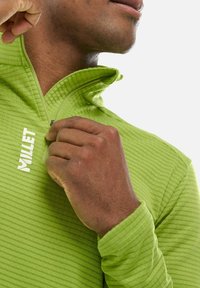 Haut zippé vert avec des rayures verticales texturées et un col montant. Présente un logo blanc sur la poitrine. Tissu doux et flexible.