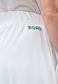 Κοντινό πλάνο ενός ατόμου που φοράει λευκά αθλητικά σορτς με πράσινο κείμενο "BORG" κοντά στη λαστιχένια μέση και μικρές μαύρες οπές αερισμού.