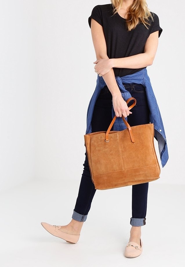 Tan suede tote bag met leren handvatten, groot rechthoekig ontwerp en gestikte details. Draag gedragen met donkere jeans en een blauwe shirt om de taille geknoopt.