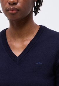 Lähivaade inimesest, kes kannab tumesinist V-kaelusega kampsunit, millel on vasakul rinnal väike tikitud logo, taust on ühevärviline.