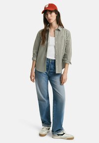 Striped button-up shirt in green and white, white tank top, light blue jeans, and cream sneakers with black accents. Red cap with logo.