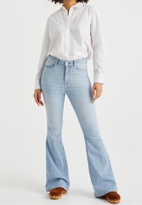 High-waisted light blue flared jeans with front pockets, paired with a white button-up shirt and brown clogs. Denim has a smooth texture.