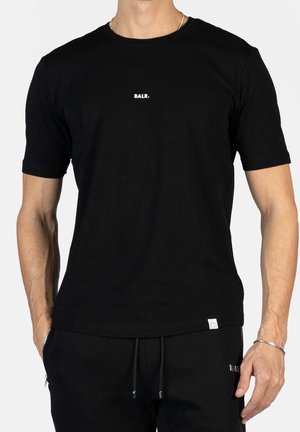 Man draagt een zwart T-shirt met korte mouwen met de tekst "BALR." op de borst en een zwarte broek, staand met handen ontspannen langs het lichaam.