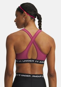 Bordeaux sportbeha met gekruiste banden, met een brede zwarte Under Armour band aan de onderkant. Glad textuur, atletisch ontwerp.