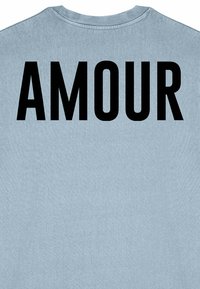 Šviesiai mėlynas medvilninis marškinėliai su ryškiu juodu tekstu, kuris nurodo "AMOUR" ant nugaros. Paprastas dizainas su apvalia iškirpte.