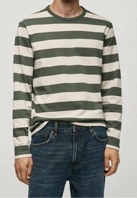 Person, der ein langärmliges Shirt mit grünen und weißen horizontalen Streifen und blaue Jeans trägt, steht mit entspannten Armen an den Seiten.