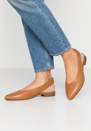 Kobieta w niebieskich dżinsach i beżowych skórzanych balerinach typu slingback na niskim obcasie, stojąca na białej podłodze na tle jednolitego tła.