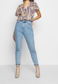 Béžový top s krátkým rukávem, na kterém jsou modré a černé vlnité vzory, v kombinaci se světle modrými džínami s vysokým pasem a béžovými vysokými botami na kotníku.