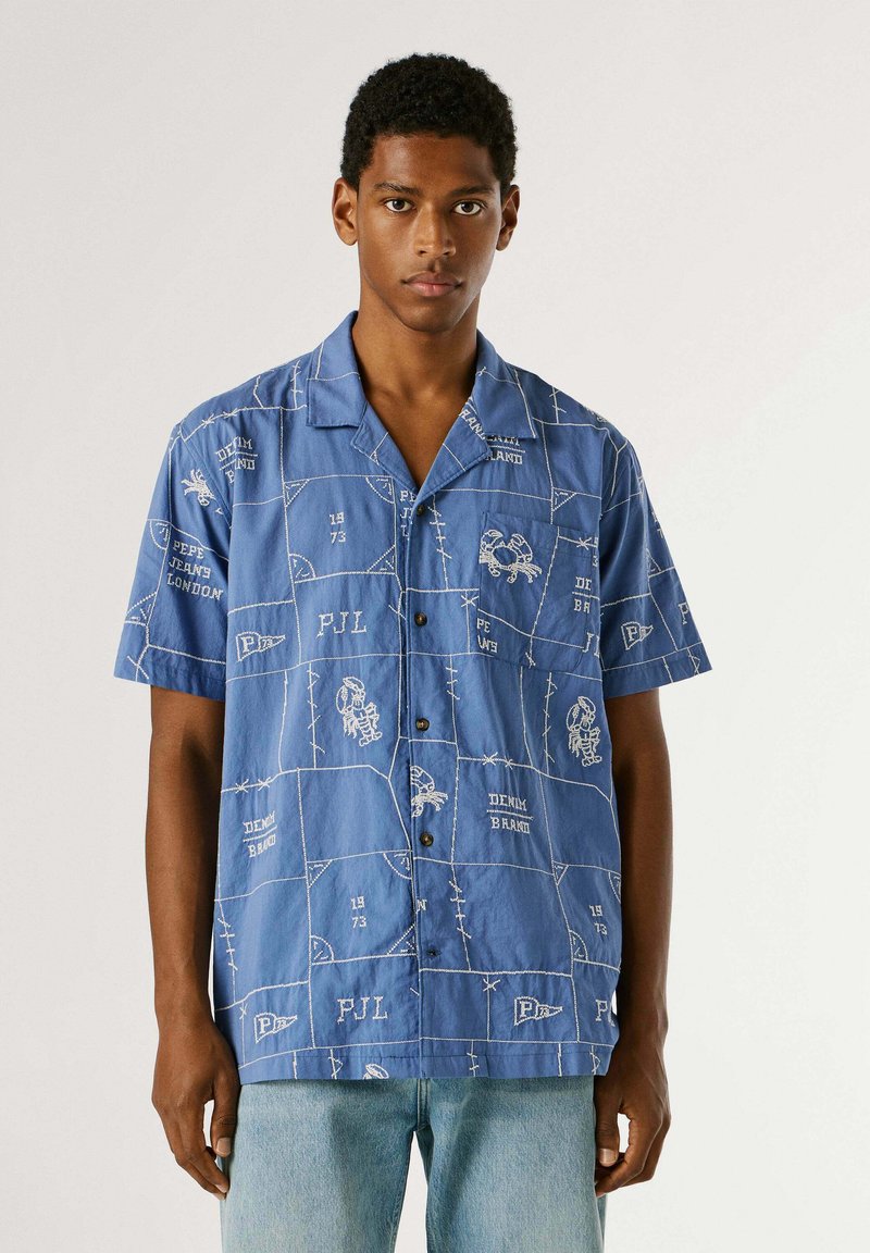 Junger Mann, der ein blaues kurzärmeliges Hemd mit weißen Stickmustern und hellblauen Jeans trägt, steht vor einem einfarbigen Hintergrund.