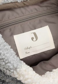 Intérieur en polaire avec une étiquette beige comportant les sections "J'appartiens à" et "Téléphone". L'étiquette présente un logo stylisé en "J" de couleur grise.
