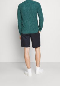 Grön texturerad tröja, marinblå shorts och vita höga sneakers. Tröjan har ribbad krage och muddar. Shortsen är knälånga.
