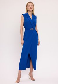 Niebieska sukienka bez rękawów z krojem kopertowym, z złotym paskiem w talii i rozcięciem z przodu, wykonana z gładkiego materiału.