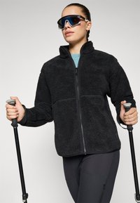 Zwarte fleece jas met volle rits en hoge kraag, gecombineerd met zwarte sportlegging. Model houdt trekkingstokken vast en draagt een donkere vizierzonnebril.
