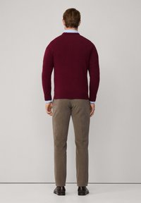 Pull en maille bordeaux, coupe ajustée, col rond, poignets côtelés, associé à un pantalon sur mesure marron clair et des chaussures foncées. Fond uni.