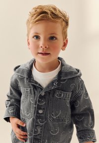 Giacca di jeans con tessuto grigio, dotata di cappuccio e motivi con faccine sorridenti, bottoni a pressione e due tasche frontali. Indossata sopra una maglietta bianca.