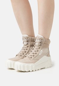 Beige Fila sneakers met hoge schacht en dikke witte chunky zolen, gedragen op blote benen, vooraanzicht tegen een effen lichte achtergrond.