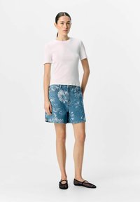 Top corto a maniche corte bianco a coste, pantaloni corti in denim blu con motivo floreale e ballerine nere in rete. Modella in piedi contro uno sfondo neutro.