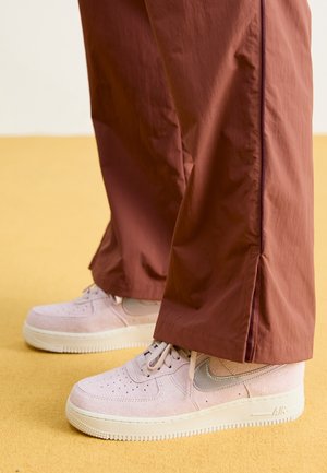 Personne portant des baskets Nike Air Force 1 en daim rose clair associées à un pantalon marron ample, debout sur un sol jaune.