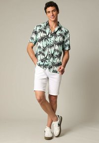 Turquoise shirt met korte mouwen met zwarte palmboompatronen, witte shorts en witte sneakers met donkere accenten. Casual zomeroutfit.