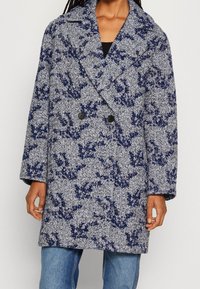 Vrouw in een middellange dubbele coltrui met een marineblauw en wit abstract patroon over blauwe jeans.