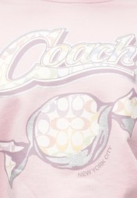 Ανοιχτό ροζ μπλουζάκι με υφασμένο γραφικό ζαχαρωτού και τη λέξη «Coach» σε μεταλλικό ασημί, με την επιγραφή «New York City» από κάτω σε γκρι.