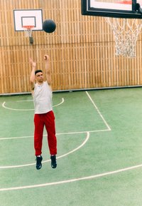 Muški sportaš u bezrukavnoj sivoj majici i crvenim hlačama skače da bi bacio crne košarkaške lopte prema obruču na zelenom terenu s bijelim oznakama.