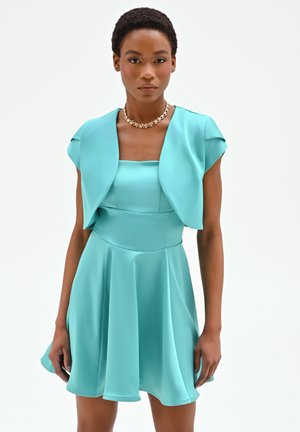 Donna con un abito turchese senza maniche con coprispalle a mantella e una collana d'oro a catena, in piedi davanti a uno sfondo bianco.