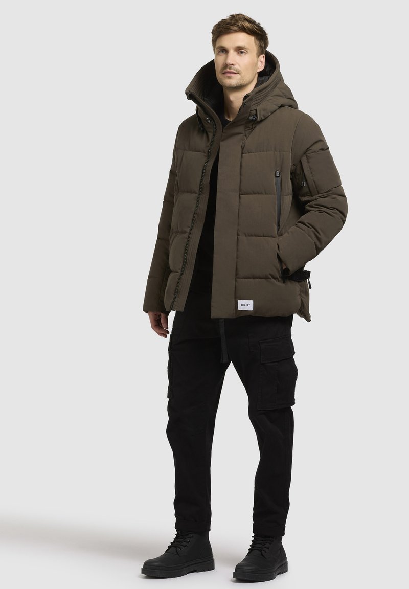 Olivengrüne Pufferjacke mit Kapuze, Reißverschlusstaschen und gestepptem Muster, kombiniert mit schwarzen Cargo-Hosen und schwarzen Stiefeln.