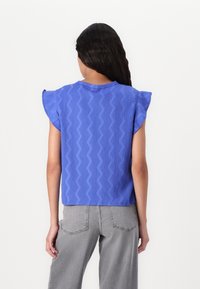 Modrá krátkosrstá blůza s texturovaným cik-cak vzorem, s kulatým výstřihem a volným střihem. V kombinaci se šedými kalhotami s vysokým pasem.