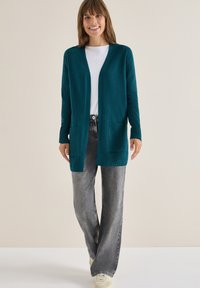 Tyrkysový pletený cardigan s dlouhými rukávy, otevřenou přední částí, dvěma předními kapsami, spárovaný se šedými širokými džíny a bílými teniskami.