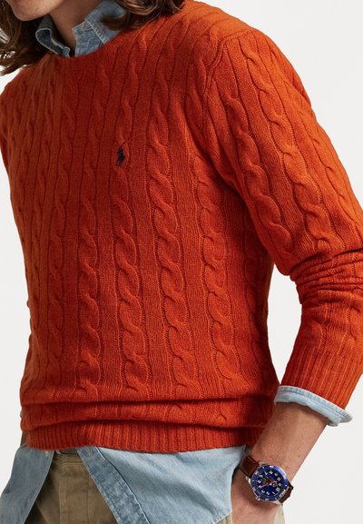 Pull en maille torsadée orange avec des poignets et un ourlet côtelés, porté sur une chemise à col bleu clair. Montre bleue avec un bracelet en cuir visible.