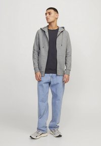 Gray zip-up hoodie with drawstrings, worn over a dark t-shirt. Paired with light blue jeans and gray athletic shoes. Casual fit, cotton blend.
