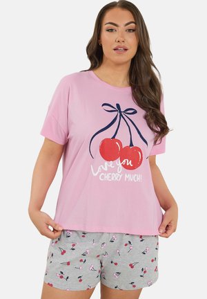 Kvinde iført lyserød T-shirt med stort kirsebærgrafik og teksten "Elsker dig kirsebær meget!", kombineret med grå shorts med små kirsebærmønstre.