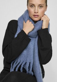 Sciarpa lavorata a maglia blu chiaro con estremità frangi, realizzata in un materiale morbido, indossata intorno al collo sopra una maglietta nera.