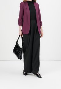 Blazer bordeaux, haut noir côtelé, pantalon large noir et chaussures noires vernies. Porte un sac à main noir mousseline avec un détail de boucle à l'avant.